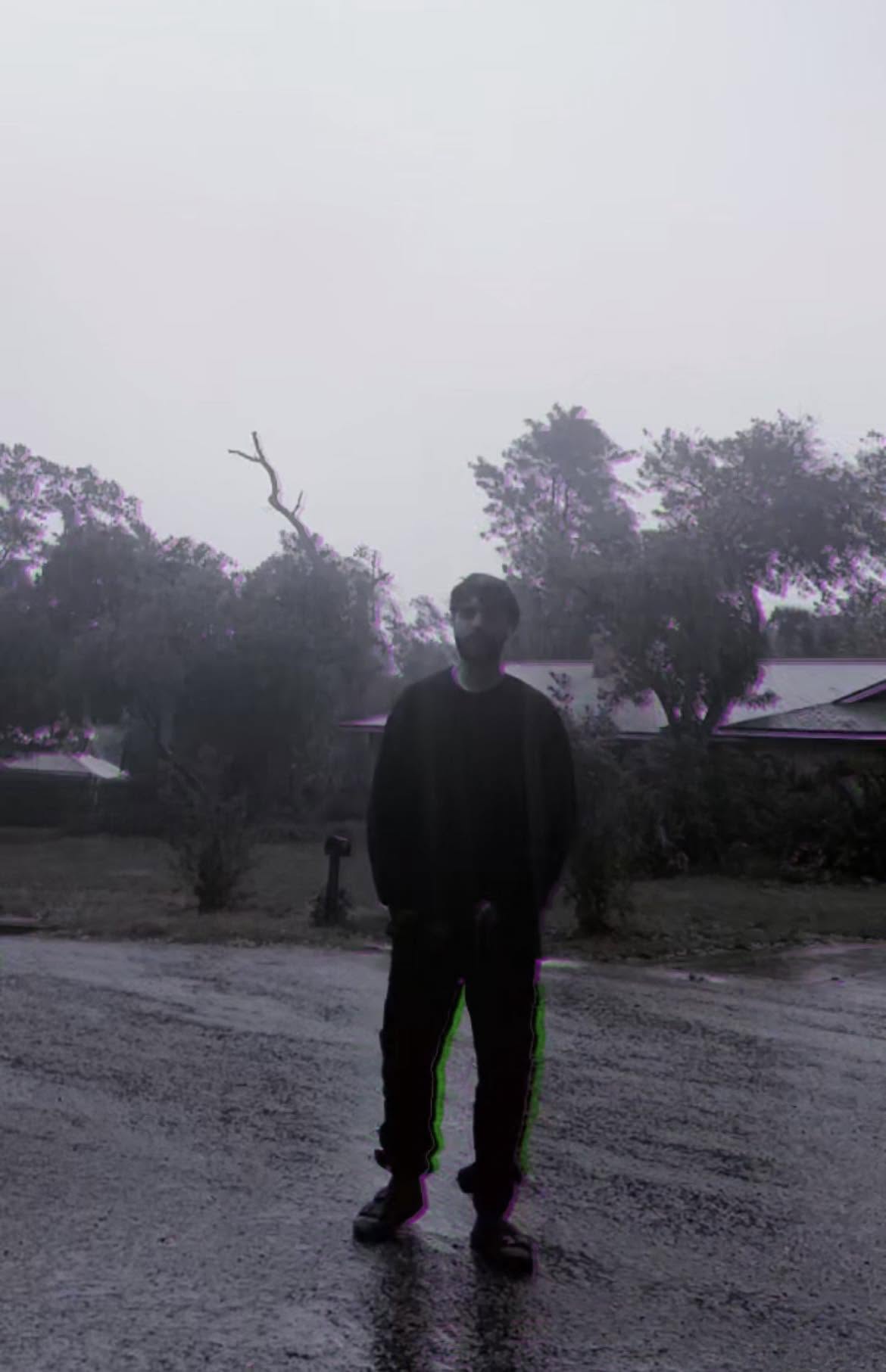 Elias standing alone in a rain-soaked street, silhouetted against trees and houses beneath a grey sky