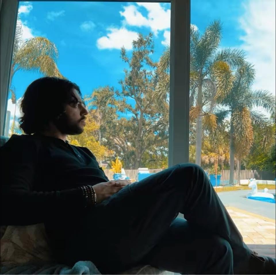 Elias sitting quietly by a large window, looking out at palm trees in reflection